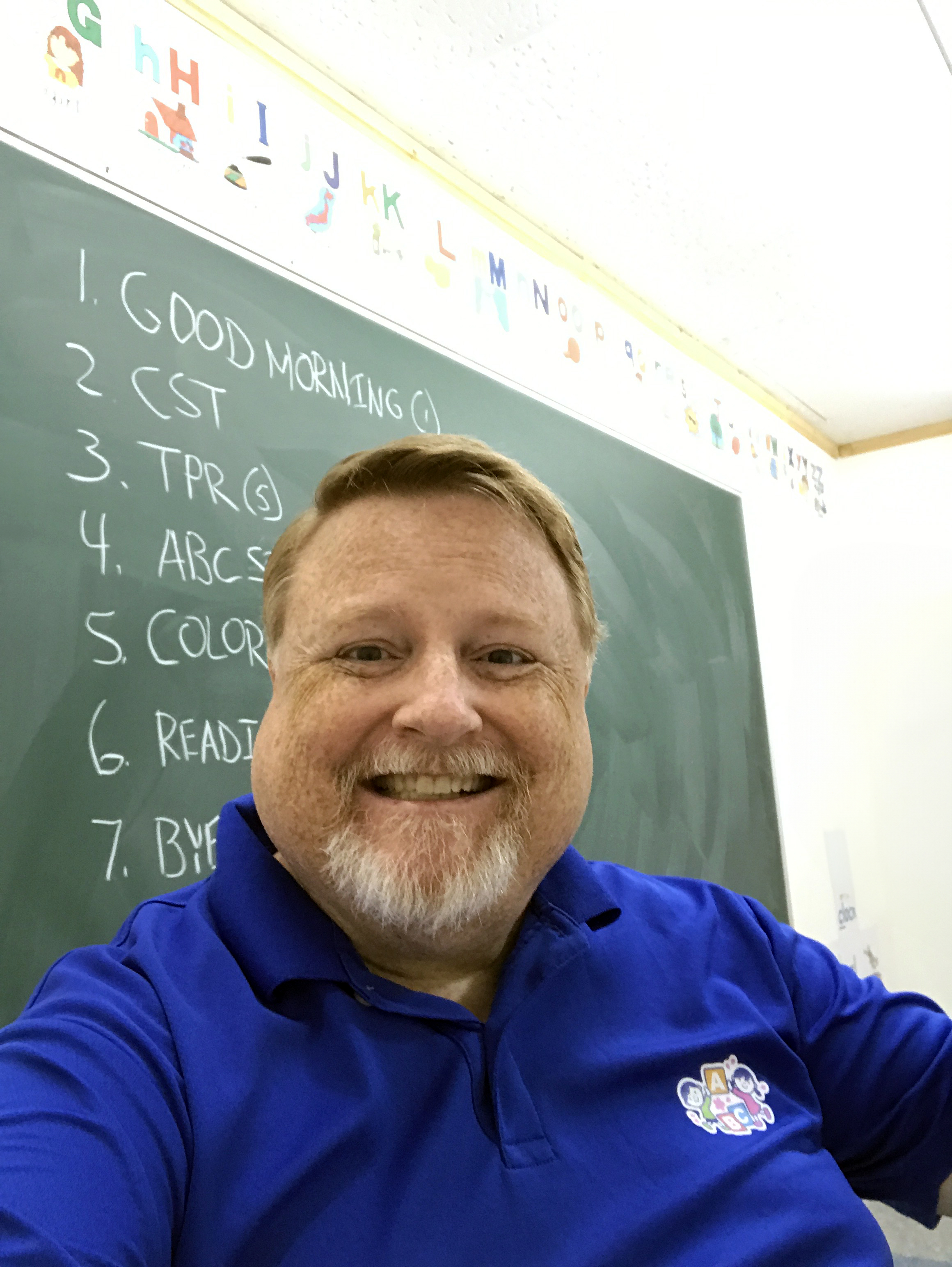 Professor Patrick in front of a green chalkboard with an English class schedule written on it.